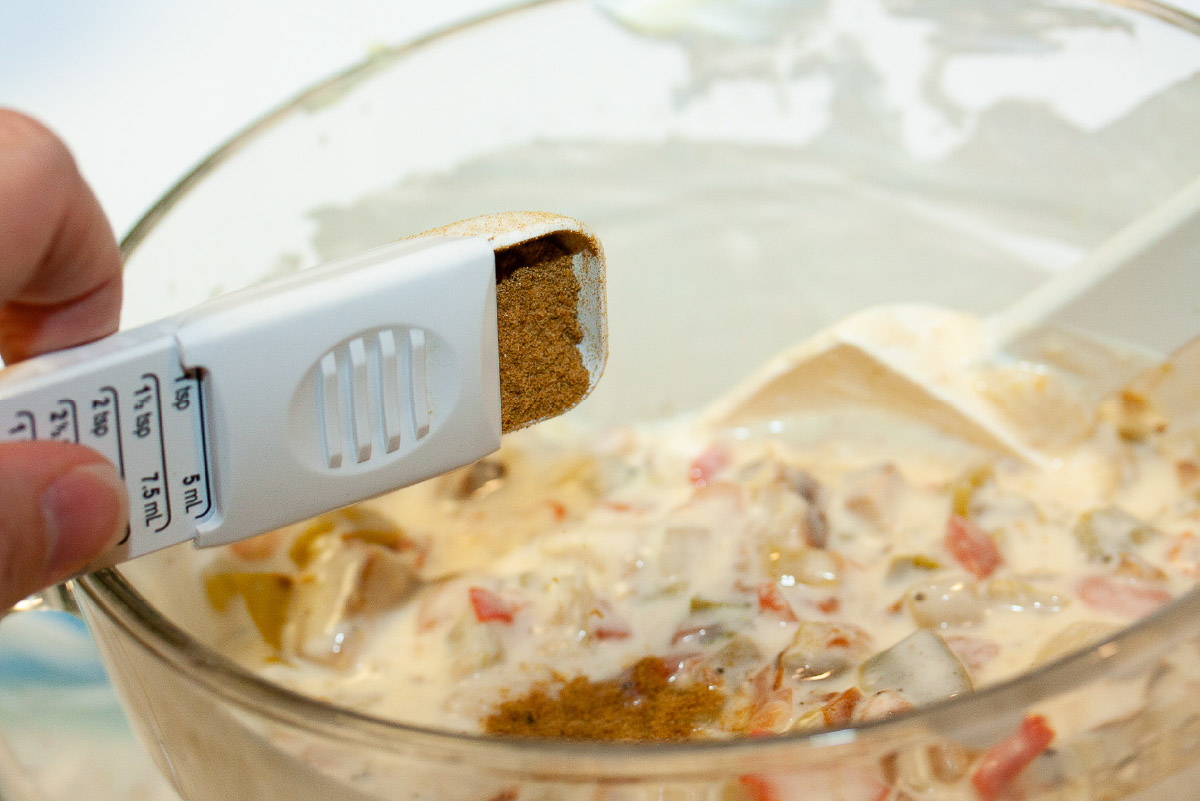 Cumin being dumped into a bowl of white cream sauce with chicken and peppers