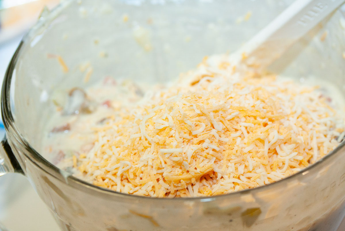 Mixing shredded Mexican blend cheese into a large glass bowl with white enchilada filling