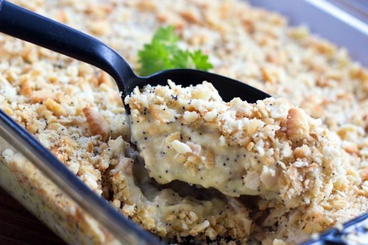 A black spoon is scooping Poppy Seed Chicken Casserole out of a clear glass casserole dish. 