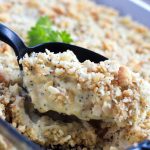 A black spoon is scooping Poppy Seed Chicken Casserole out of a clear glass casserole dish.