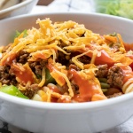 Bowl of seasoned ground beef, spiral pasta, lettuce, halved tomatoes, nacho cheese Doritos, and French salad dressing with shredded cheddar cheese on top