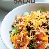 Bowl of seasoned ground beef, spiral pasta, lettuce, halved tomatoes, nacho cheese Doritos, and French salad dressing with shredded cheddar cheese on top with text Taco Mac Salad Add Salt & Serve