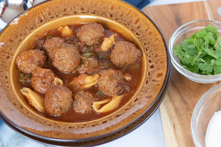 Photo is a brown soup bowl with meatball soup, beside a cutting board. On the cutting board are two clear glass bowls with parsley and sour cream.