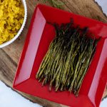 grilled asparagus on a red serving plate placed on a wood serving tray with a bowl of yellow rice and a tea towel to the side