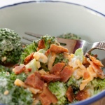 Broccoli, shredded cheddar cheese, bacon bits, and mayonnaise in a bowl with a fork.