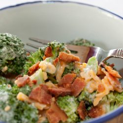 Broccoli, shredded cheddar cheese, bacon bits, and mayonnaise in a bowl with a fork.