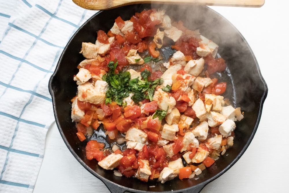 Cast iron skillet cooking chopped chicken, diced tomatoes, and fresh basil
