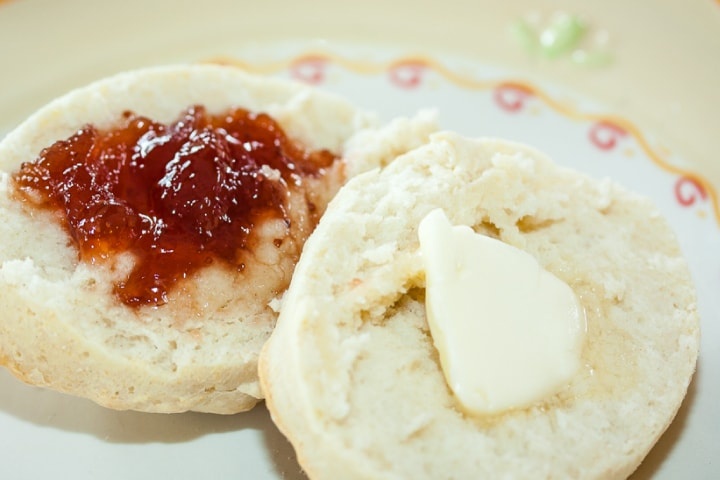 homemade-buttermilk-biscuits-post-horiz-min Biscuit cut in half with butter on one half and fruit preserves on the other half on a yellow and white plate