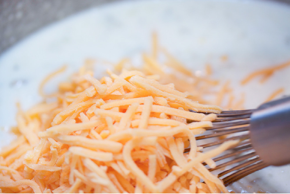 A whisk mixing shredded cheddar cheese into a bowl of baked potato soup.