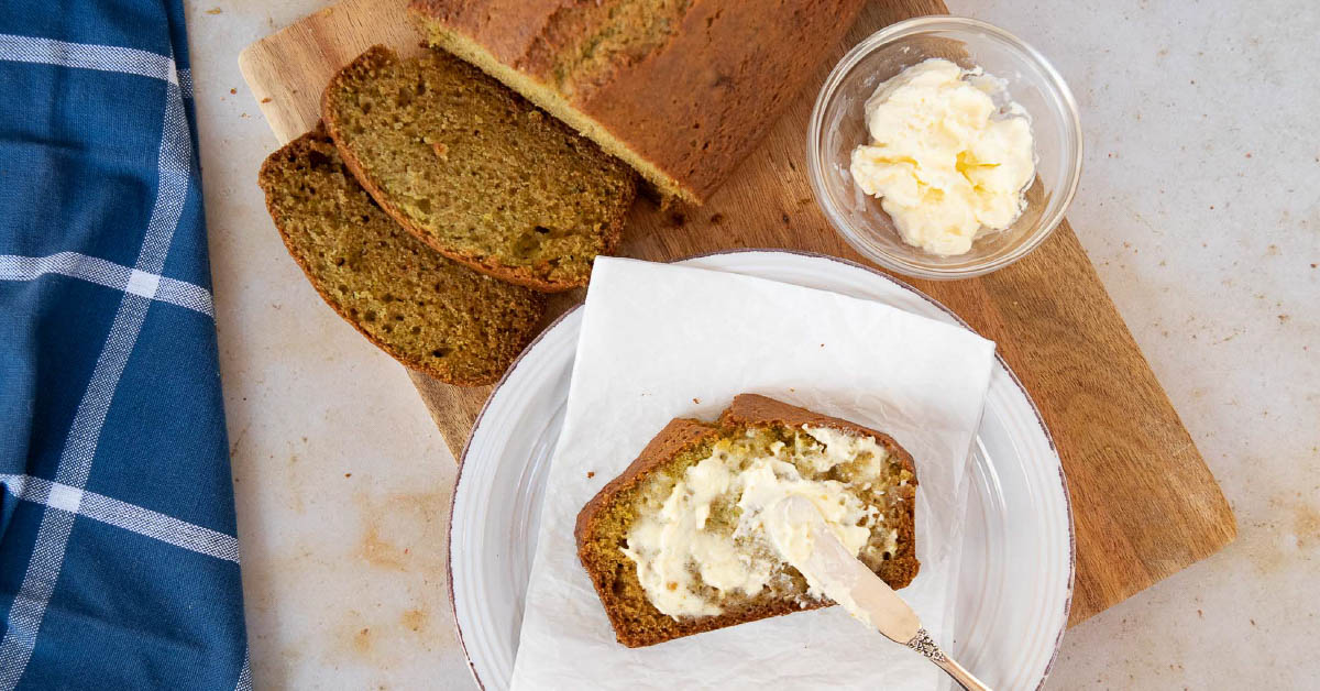 Overhead shot of a loaf of zucchini bread with a slice being spread with clotted cream