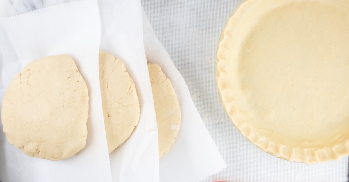 Overhead image of unbaked pie crust in a pie pan to the right and thick circles of pie crust dough stacked with parchment paper in between ready for the freezer