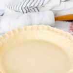 Unbaked pie crust in a pie pan with a marble rolling pin and flour sack towel in the background