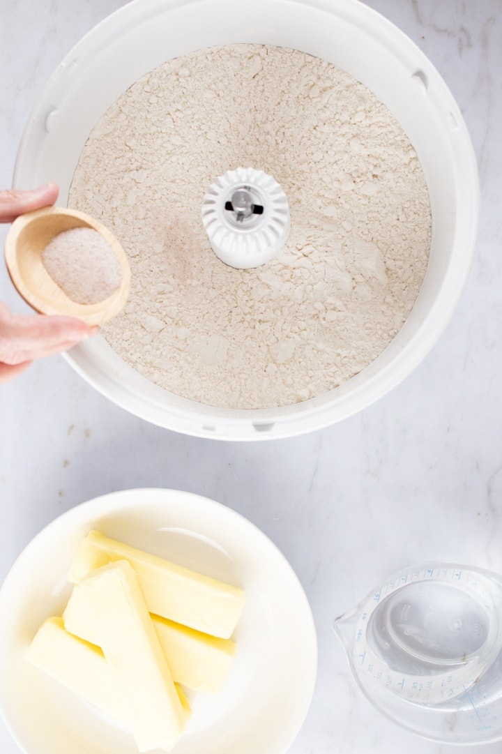 Overhead shot of flour in a mixer with salt being added from a small wooden bowl and containers of butter and water to the side