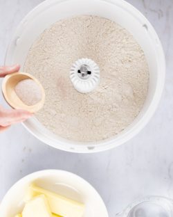 Overhead shot of flour in a mixer with salt being added from a small wooden bowl and containers of butter and water to the side