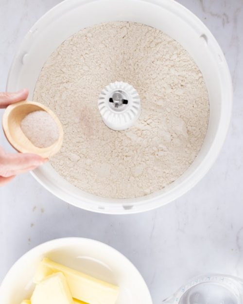 Overhead shot of flour in a mixer with salt being added from a small wooden bowl and containers of butter and water to the side