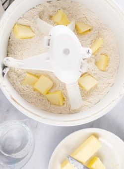 Overhead shot of flour in a mixer with chopped butter added throughout, a bowl with two sticks of butter to the side, and a measuring cup with 1-½ cups water to the side