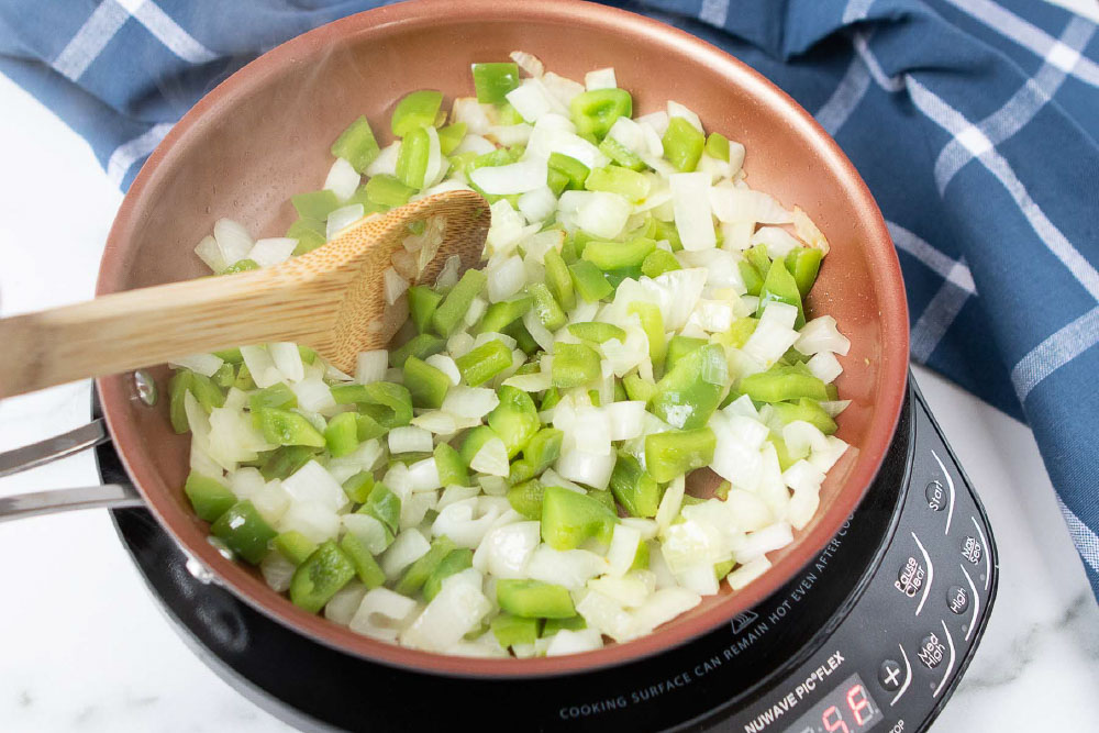 Chopped onion and green bell pepper in a fry pan being stirred with a wooden spoon