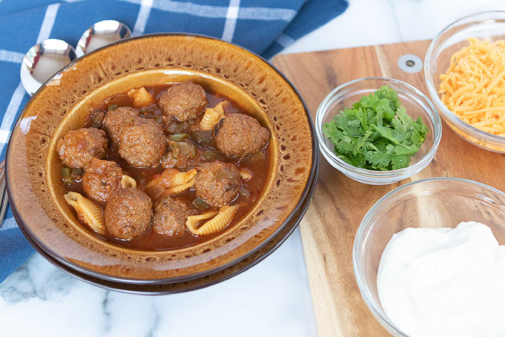 Overhead shot of albondigas soup (Mexican meatball soup) in a brown wide-rim bowl with bowls of sour cream, grated cheddar cheese, and fresh cilantro to the side