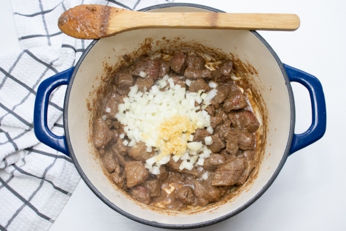 Beef stew in a pot with chopped raw onions and garlic added