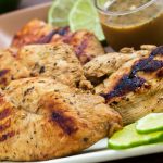 Grilled chicken on a square plate, garnished with lime slices. A small clear glass cup of Dijon basil lime dipping sauce is on the plate.