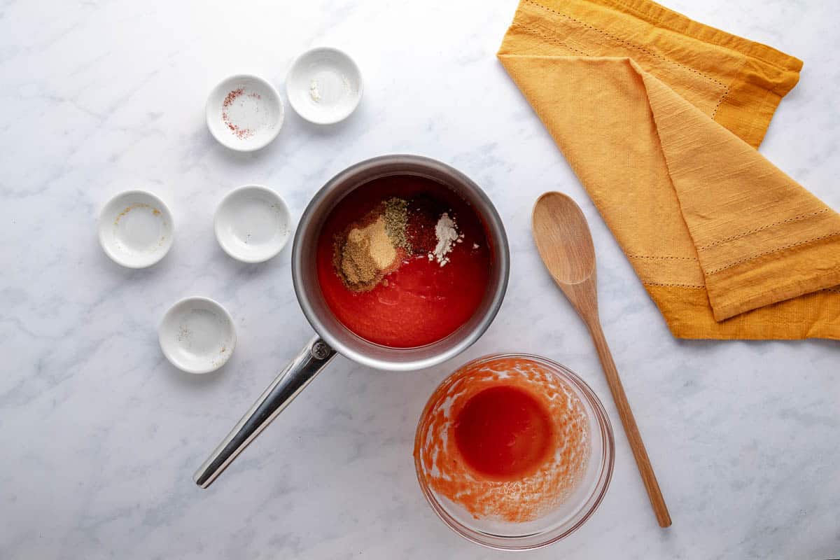 Tomato sauce, chili powder, ground cumin, onion powder, garlic powder, and dried oregano in a saucepan with a wooden spoon and a tea towel beside it on the counter.
