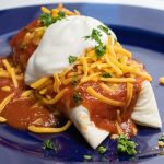 Blue plate with a burrito covered in red sauce and garnished with cheese, sour cream, and cilanto