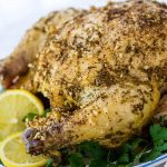 Whole roasted chicken with a dried rosemary and minced garlic rub on a white plate, garnished with lemon slices and parsley.