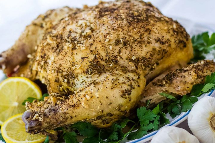 Whole roasted chicken with a dried rosemary and minced garlic rub on a white plate, garnished with lemon slices and parsley. There are two bulbs of garlic beside the plate in the bottom right corner.