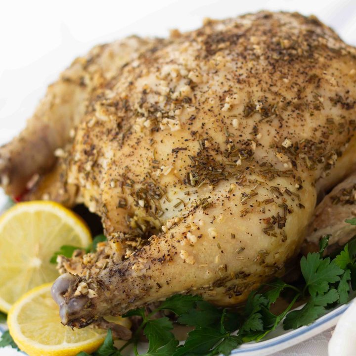 Whole roasted chicken with a dried rosemary and minced garlic rub on a white plate, garnished with lemon slices and parsley.
