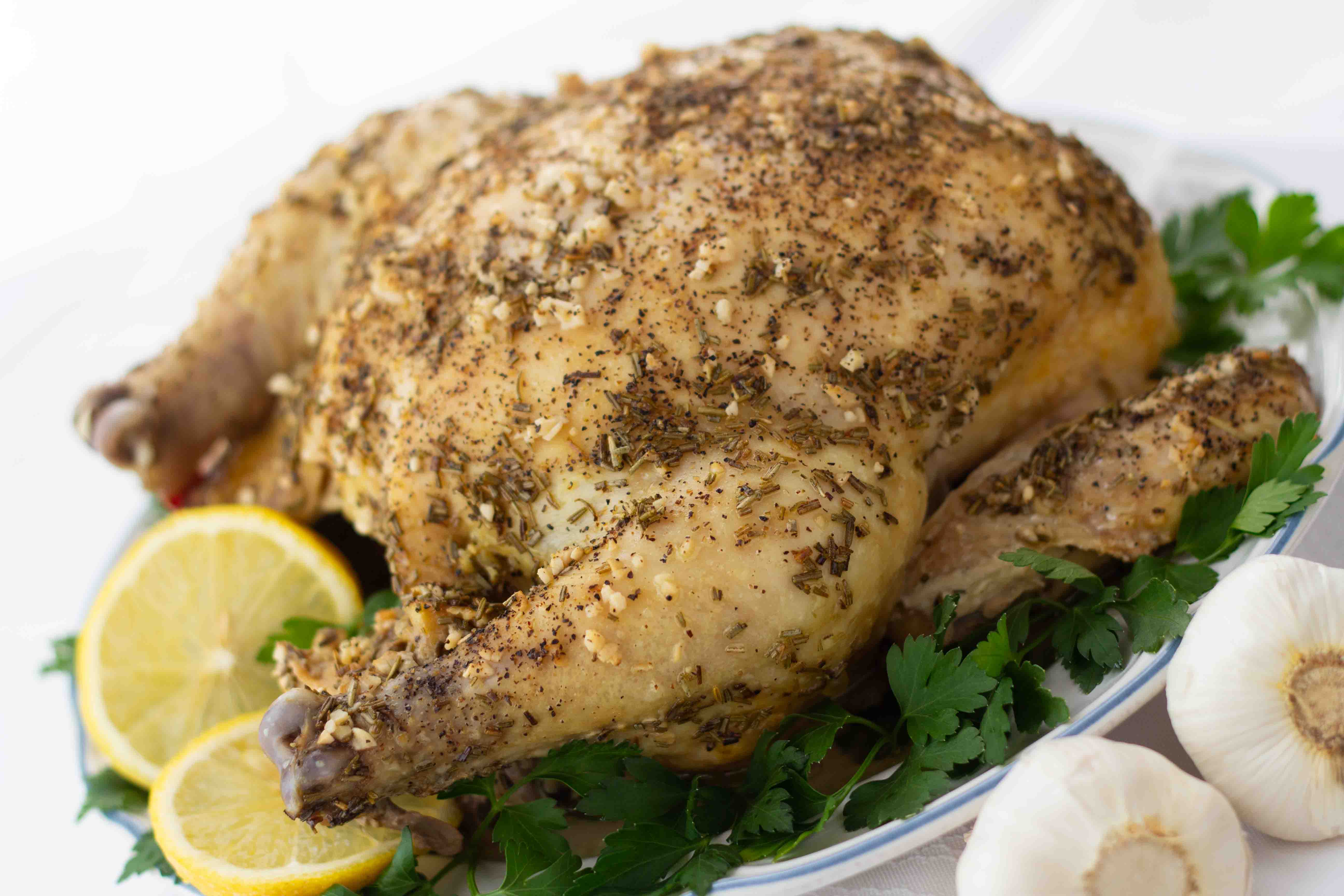 Greek Chicken Whole roasted chicken with a dried rosemary and minced garlic rub on a white plate, garnished with lemon slices and parsley. There are two bulbs of garlic beside the plate in the bottom right corner.