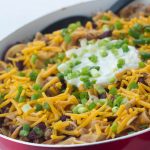 Kidney beans, ground beef, and egg noodles topped with shredded cheddar cheese, sour cream, and chopped green onions, in a skillet.
