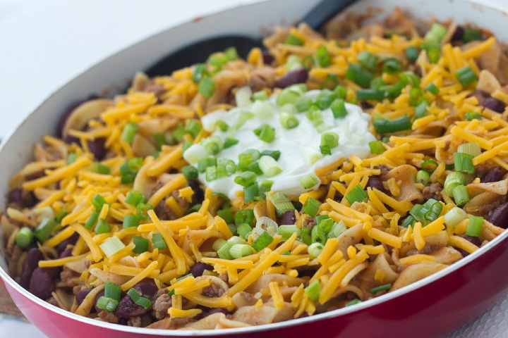 Kidney beans, ground beef, and egg noodles topped with shredded cheddar cheese, sour cream, and chopped green onions, in a skillet.
