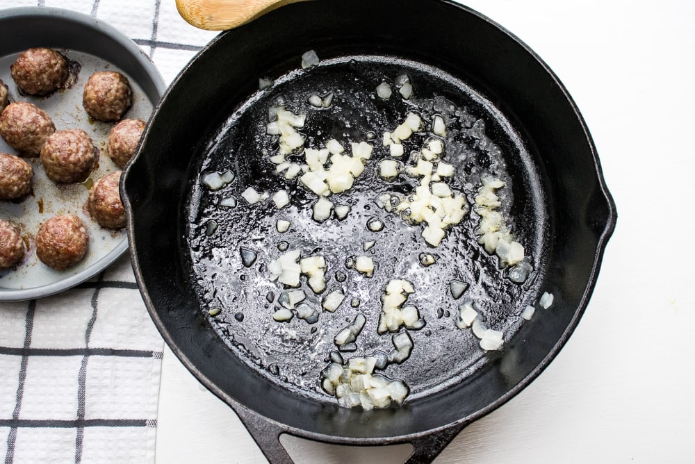 Cast iron skillet with chopped onion sautéing and meatballs in a round baking pan to the side