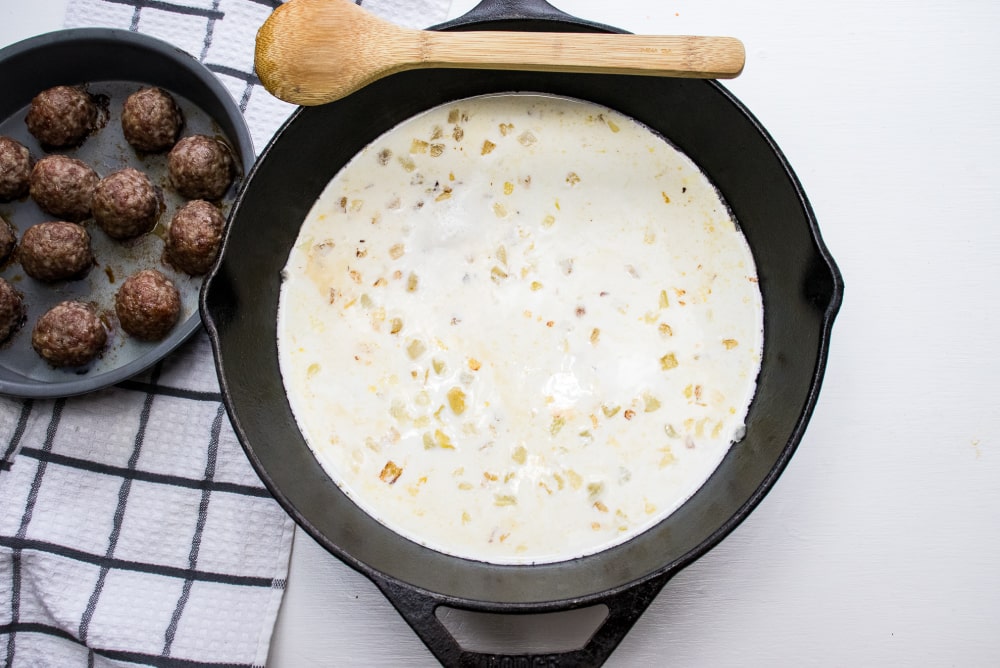 Cast iron skillet sautéed chopped onion simmering in cream and meatballs in a round baking pan to the side