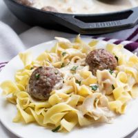 Cast iron skillet with meatballs and sauce, and two white plates with egg noodles, meatballs, and sauce.