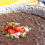 A white bowl with Black Bean Soup garnished with sour cream, diced tomatoes, diced red onion, and fresh cilantro