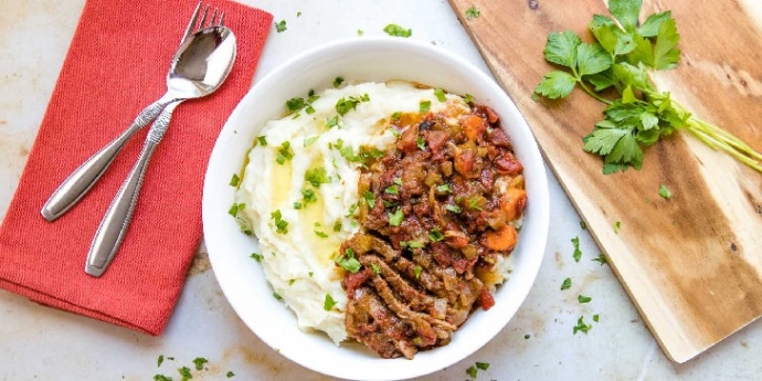 White bowl with mashed potatoes in one half and strips of beef and chopped vegetables in the other half, garnished with parsley. To the left of the bowl is a spoon and fork on a red linen napkin. To the right is a wooden cutting board with parsley on it.
