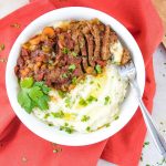 White bowl with mashed potatoes in one half and strips of beef and chopped vegetables in the other half, garnished with parsley. The bowl is on a red linen napkin and there is a fork in the bowl.