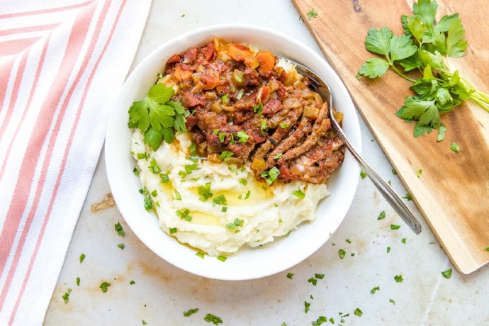 White bowl with mashed potatoes in one half and strips of beef and chopped vegetables in the other half, garnished with parsley. To the left of the bowl is a spoon and fork on a red linen napkin. To the right is a wooden cutting board with parsley on it.