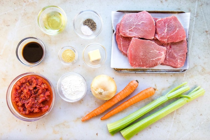 Photo is ingredients in separate clear glass bowls: diced tomatoes, Worcestershire sauce, oil, salt and pepper, dry mustard, flour, butter. There are 6 beef steaks on a metal pan. An onion, two carrots, and two stalks of celery are on the counter next to the pan.