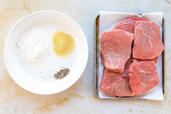 Flour, dry mustard, salt, and pepper on a white plate. 6 beef steaks on a metal pan.