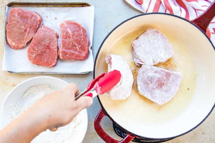 Three floured beef steaks in a red and white skillet. To the left of the skillet are three plain beef steaks on a metal pan and a white plate with the flour mixture.