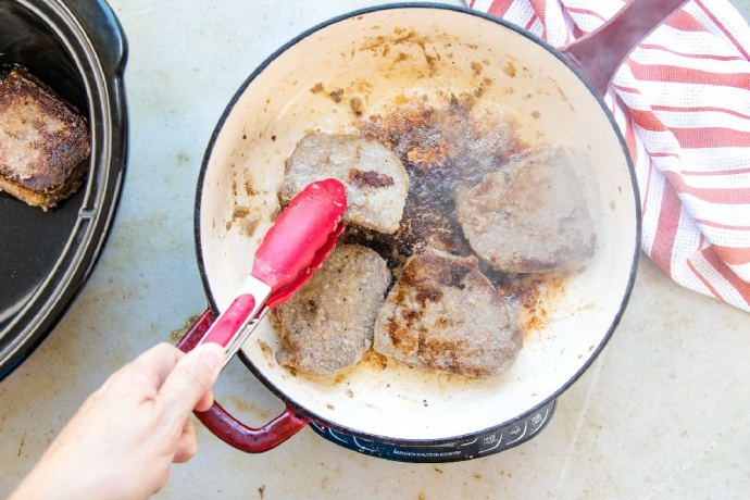 Four cooked beef steaks in a red and white skillet. To the left of the skillet is a slow cooker with one cooked steak in it. To the right is a red and white striped dish towel.
