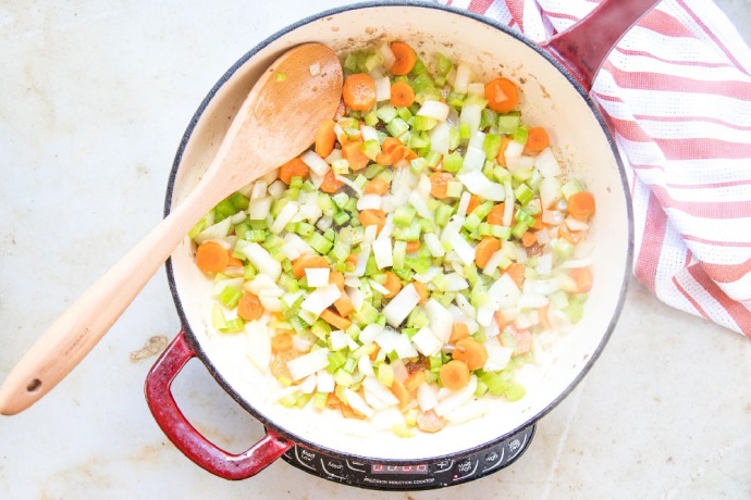 Chopped onion, carrots, and celery in a red and white skillet. There is a wooden spoon in the skillet, and a red and white striped dish towel beside the skillet.