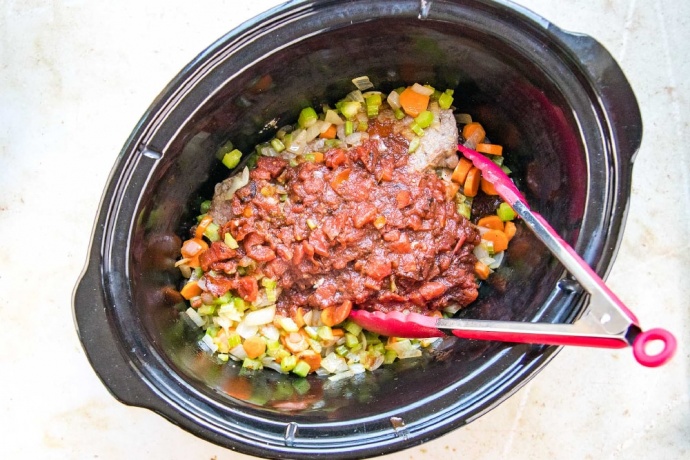 Chopped onion, carrots, and celery topped with Worcestershire sauce and tomato mixture in a slow cooker. There is a pair of red metal tongs in the slow cooker.