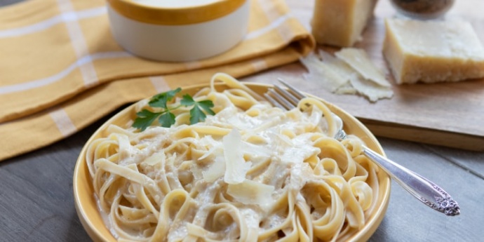 Yellow plate with fettuccine and Alfredo sauce with pot of Alfredo sauce, hunk of Parmesan cheese, and nutmeg grinder in background