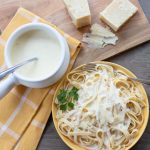 Overhead shot of yellow plate with fettuccine and Alfredo sauce with pot of Alfredo sauce, hunk of Parmesan cheese, and nutmeg grinder surrounding plate