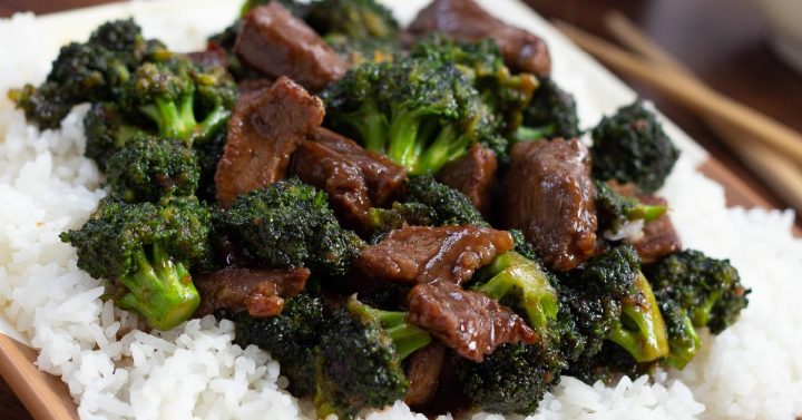 Beef slices with broccoli in a soy-based sauce served over white rice on a white plate with chopsticks to the side