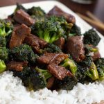 Beef slices with broccoli in a soy-based sauce served over white rice on a white plate with chopsticks to the side