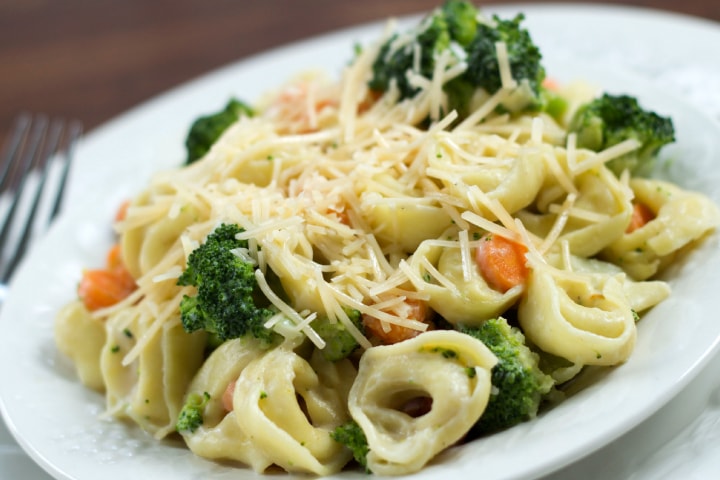 Plate of cooked tortellini and Alfredo sauce with broccoli and carrots garnished with shredded Parmesan cheese
